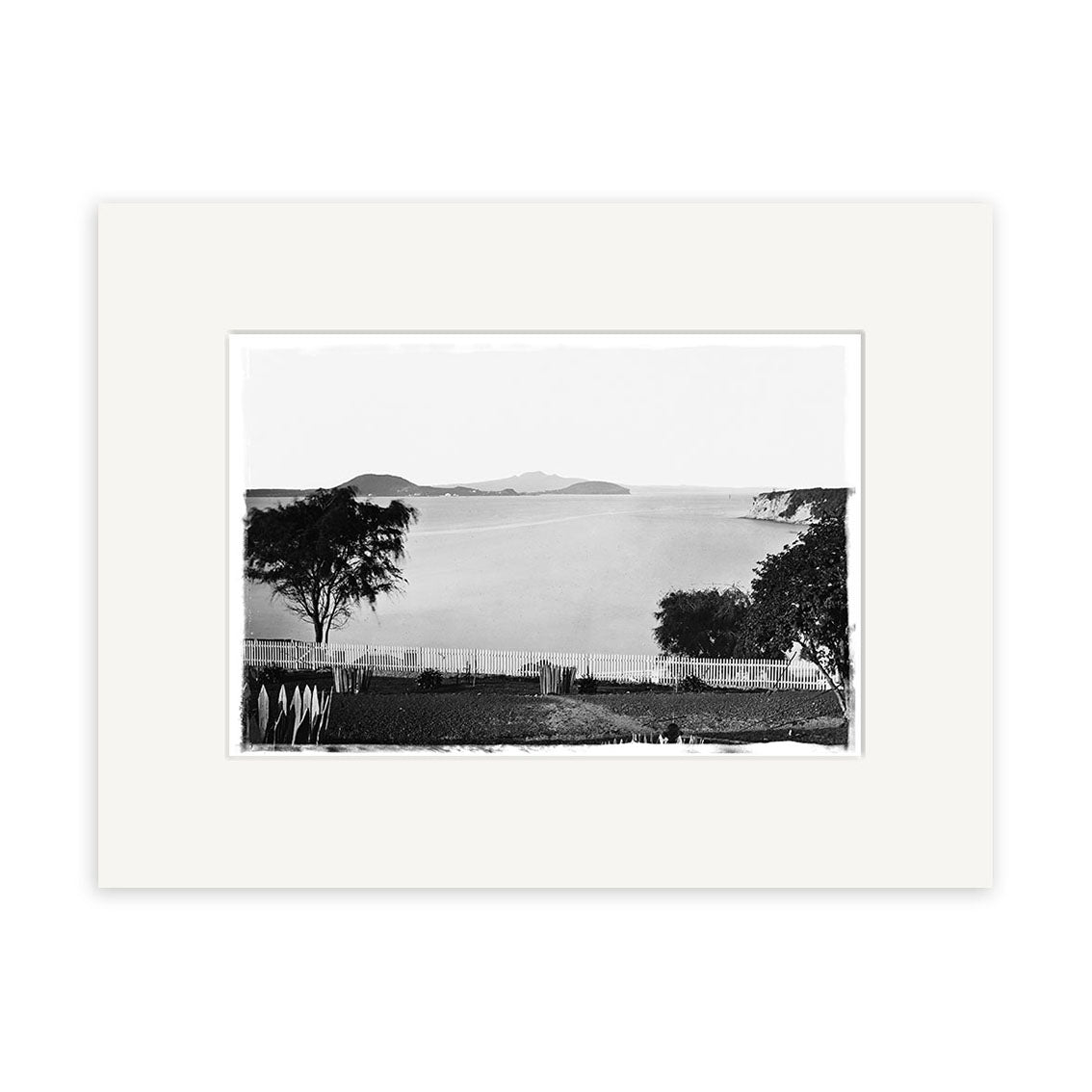 St Georges Bay 1864 - HI049 is a black and white photo of calm water with distant islands, framed by trees and a white fence. This historical print is mounted with a white mat on archival quality matte paper.