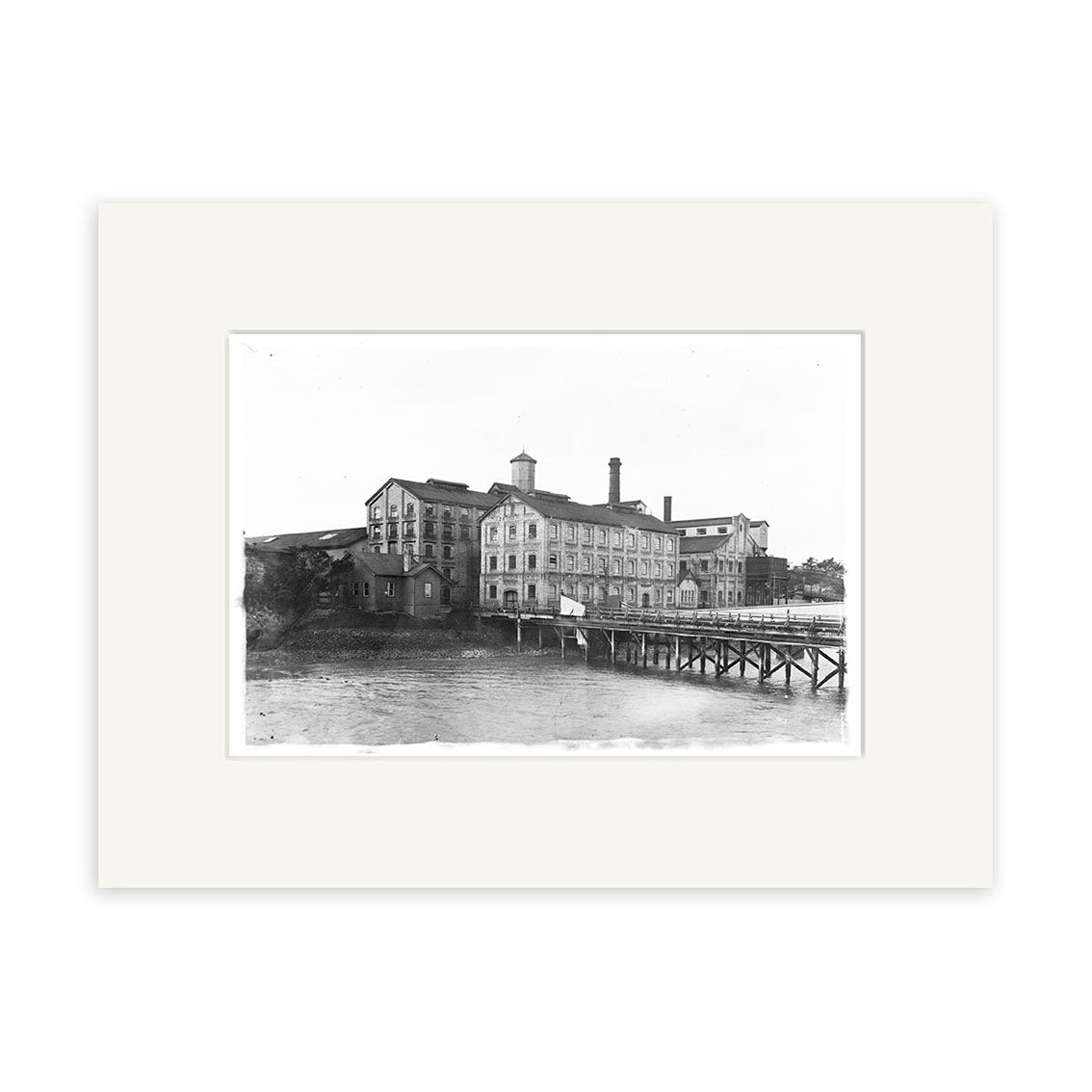 CSW 1915 - HI050: Black and white historical photographic print of a large multi-story industrial building with chimneys by water in New Zealand, seen from a wooden pier; archival quality, framed with a white mat.