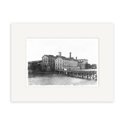 CSW 1915 - HI050: Black and white historical photographic print of a large multi-story industrial building with chimneys by water in New Zealand, seen from a wooden pier; archival quality, framed with a white mat.