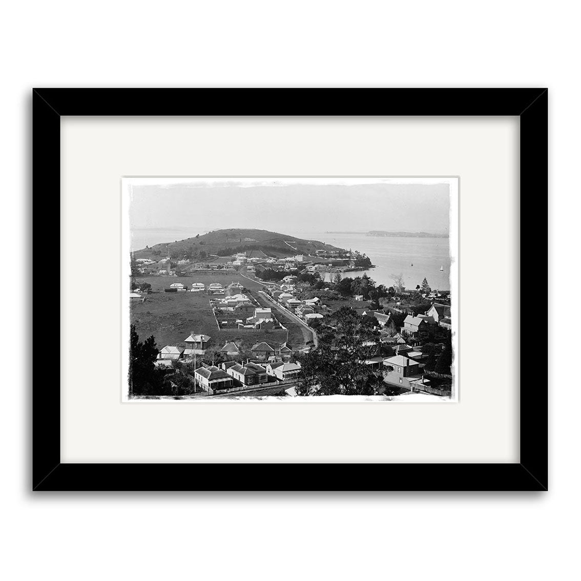 Devonport 1880 - HI058: Archival black-and-white print of a New Zealand coastal town with houses, trees, road, hill, and water. Framed with a white mat and black border.