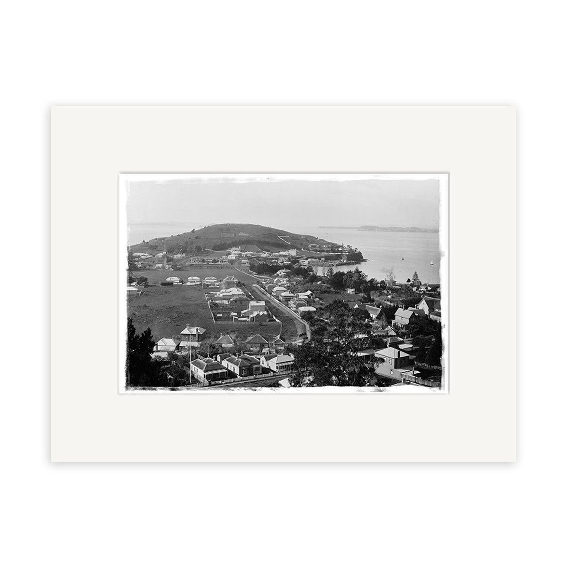 Black and white photo of Devonport 1880 - HI058, showing a coastal New Zealand town with houses, dirt roads, greenery, and a hill by the water. This historical print has a wide white mat border and is made on archival quality paper.
