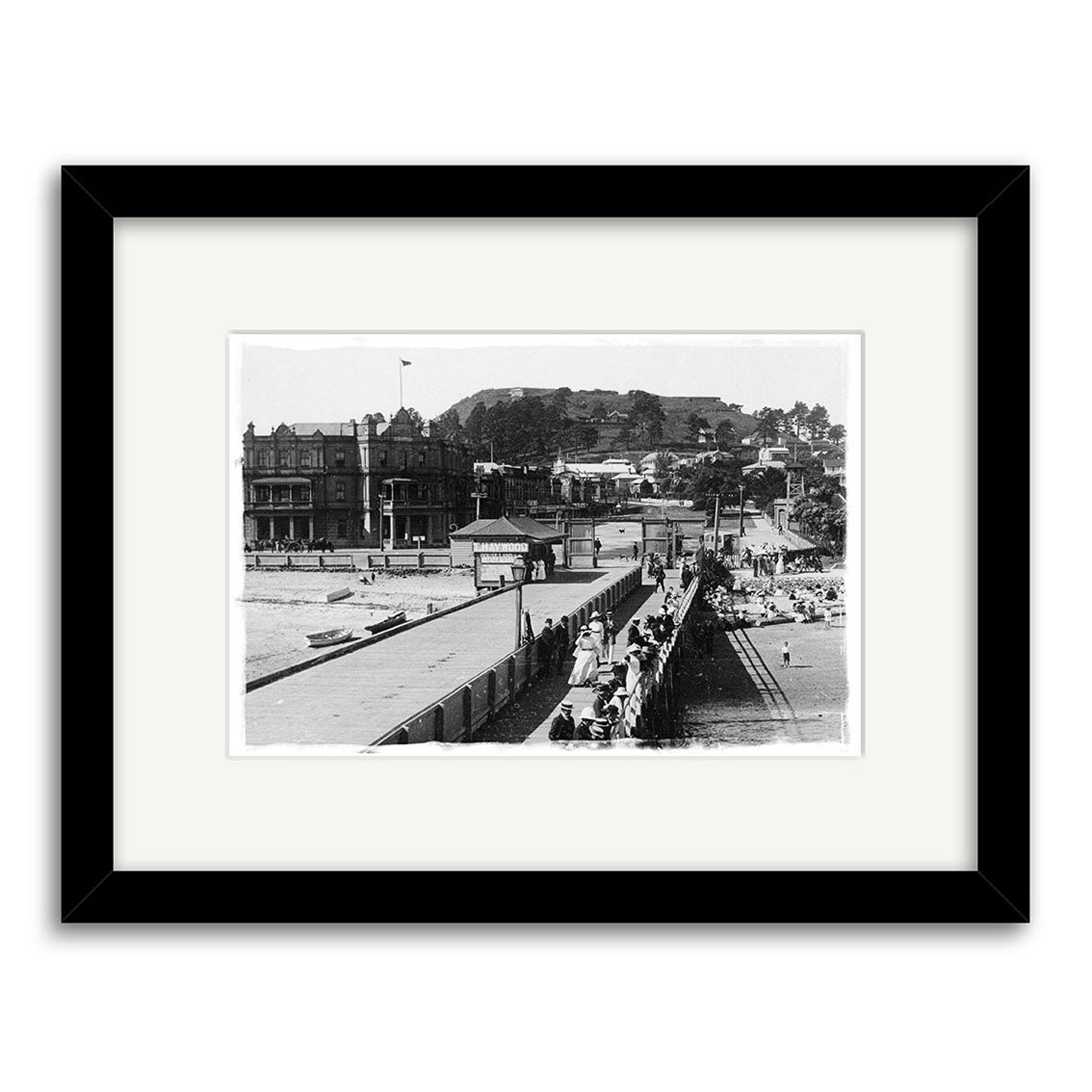 Devonport Wharf 1910 - HI059: Black and white historical photo print of a pier with people, buildings, and a hill in the background. Made in New Zealand, matted and set in a black frame.
