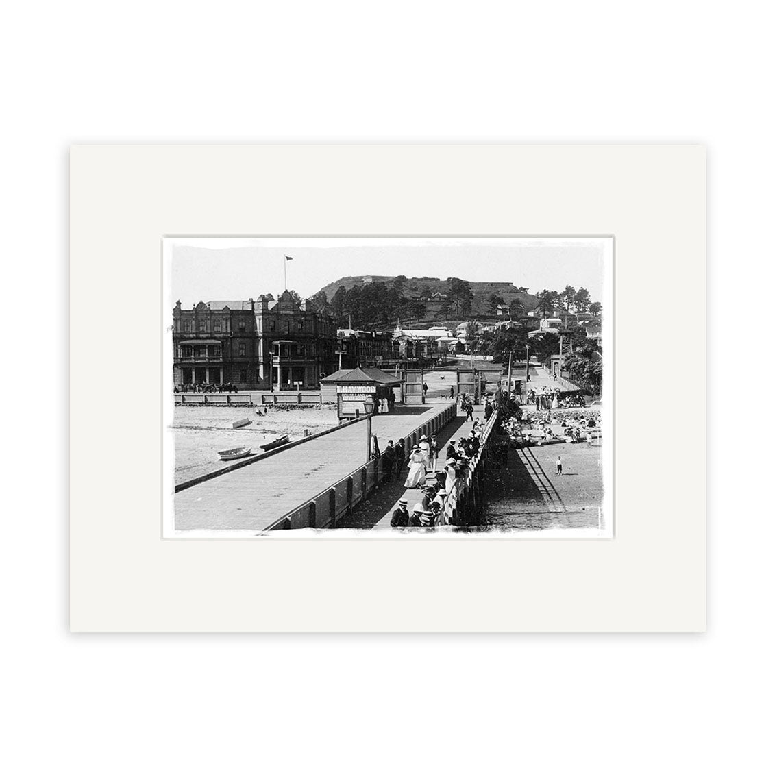 Devonport Wharf 1910 - HI059: Archival quality black and white photo of people walking on a wide pier with railings, leading to buildings and a hillside, set within white matting.