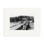 Devonport Wharf 1910 - HI059: Archival quality black and white photo of people walking on a wide pier with railings, leading to buildings and a hillside, set within white matting.