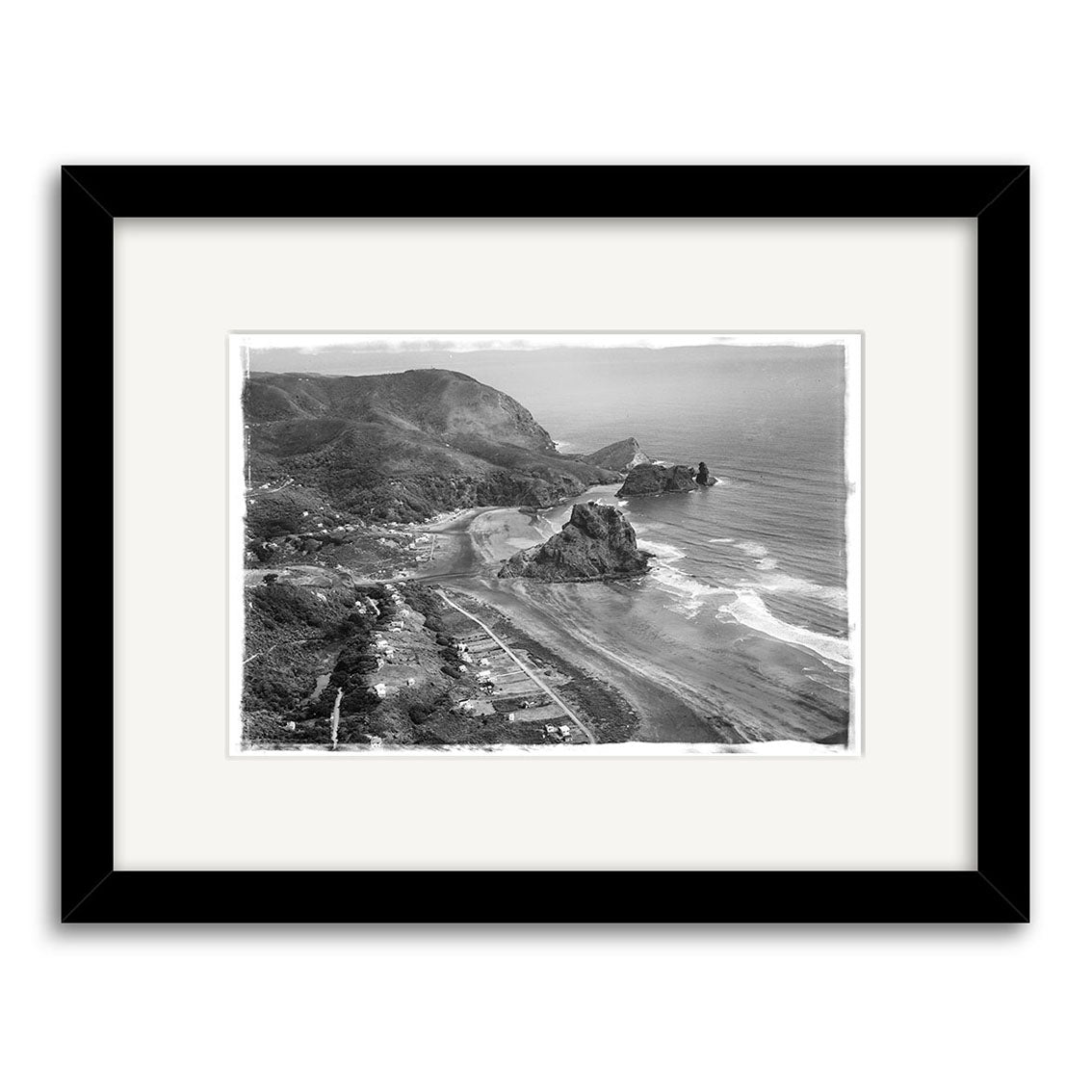 Piha 1953 - HI060: Black and white aerial photograph of a coastal landscape with beach, rocks, and hills, presented in a black frame with white mat. This historical print is expertly made on archival matte paper.