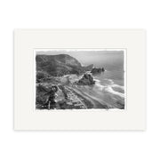 The Piha 1953 - HI060 Historical Photographic Print features a black and white aerial view of rugged cliffs, sandy beach, waves, hills, and sparse buildings. It comes bordered with a white mat for archival-quality display.