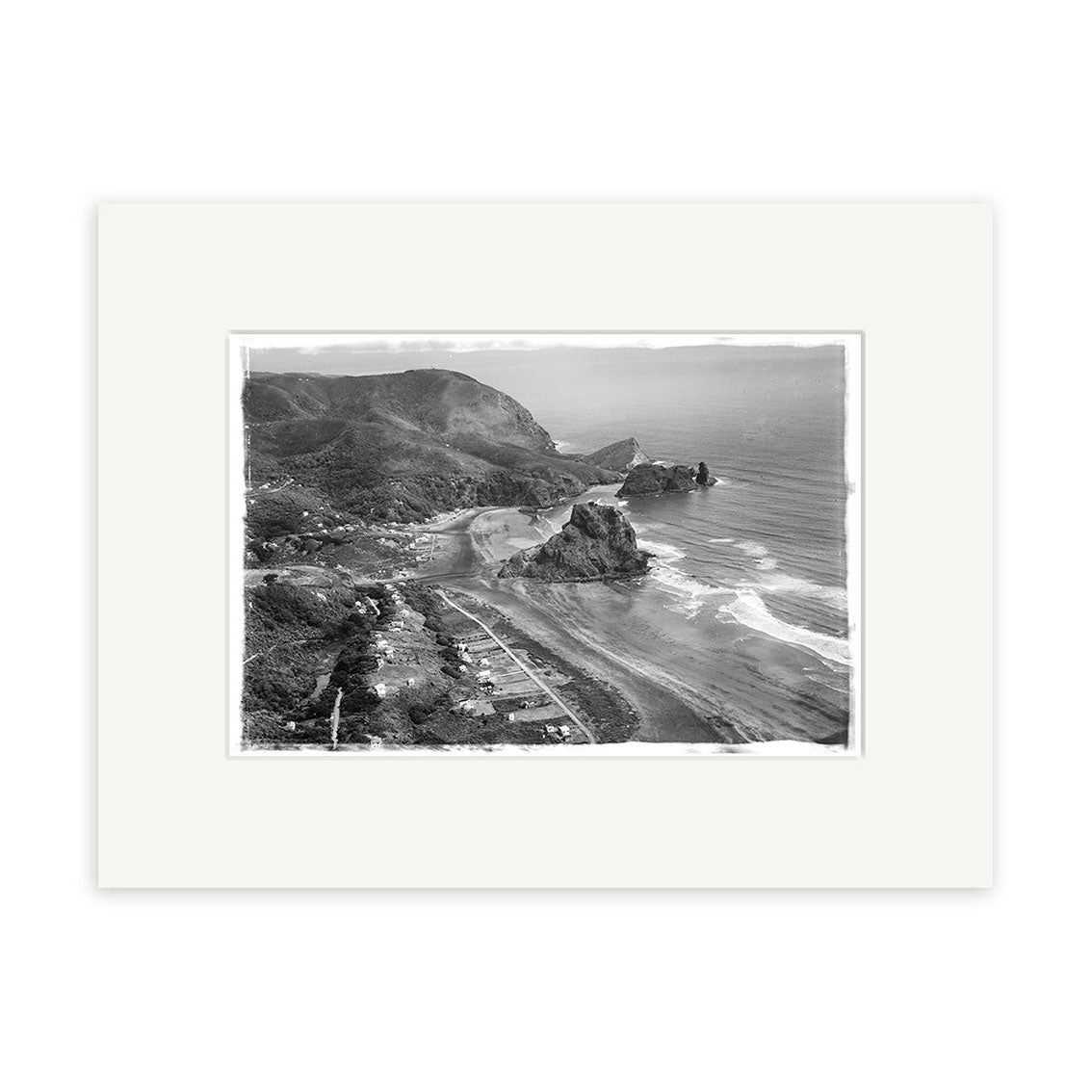 The Piha 1953 - HI060 Historical Photographic Print features a black and white aerial view of rugged cliffs, sandy beach, waves, hills, and sparse buildings. It comes bordered with a white mat for archival-quality display.