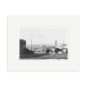 A black-and-white historical print, College Hill 1905 - HI061, depicts a New Zealand street with trolley tracks, horse-drawn carriages, utility poles, houses on both sides, and several people against a townscape backdrop.