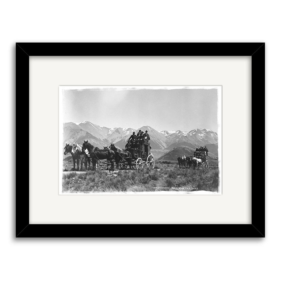 Black-and-white photo titled Christchurch Rd Cass 1900 - HI062 shows two horse-drawn stagecoaches with passengers crossing grassy land before snow-capped mountains. Historic New Zealand print, framed with white mat and black border.