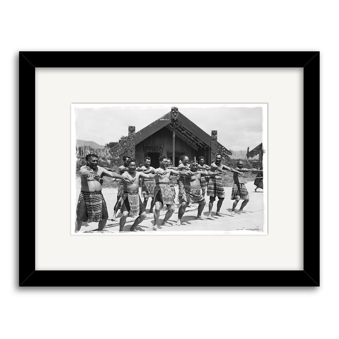The archival-quality Haka - HI063 features a black and white photo of men performing a haka in front of a Māori meeting house, beautifully framed with a white mat and black frame, capturing this powerful cultural moment in New Zealand.