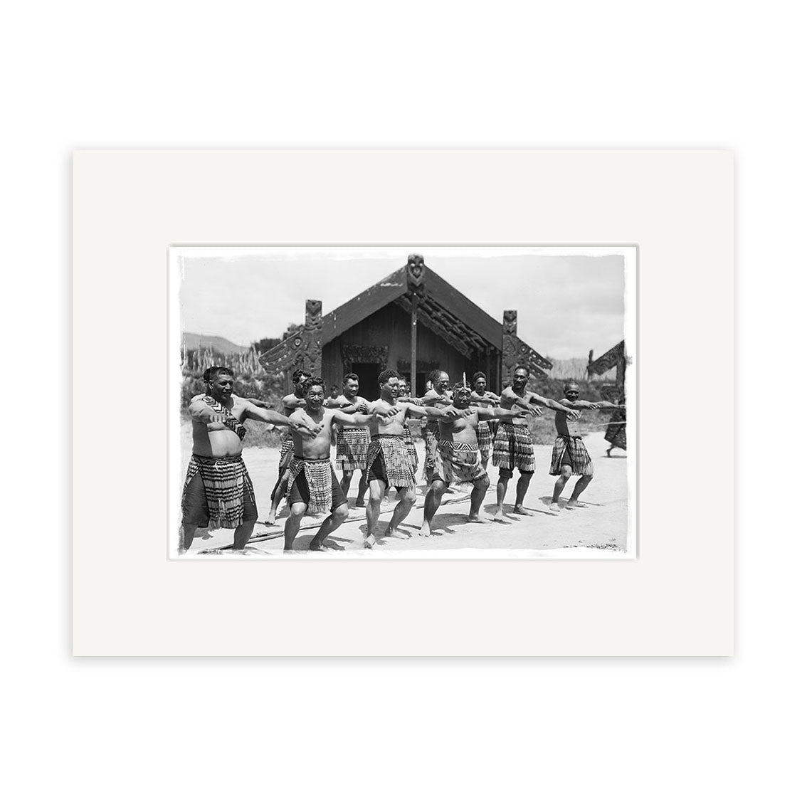 A group of Māori men in traditional dress perform a haka with arms extended before a carved meeting house on a clear New Zealand day, captured in the archival-quality photographic print Haka - HI063.