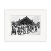 A group of Māori men in traditional dress perform a haka with arms extended before a carved meeting house on a clear New Zealand day, captured in the archival-quality photographic print Haka - HI063.