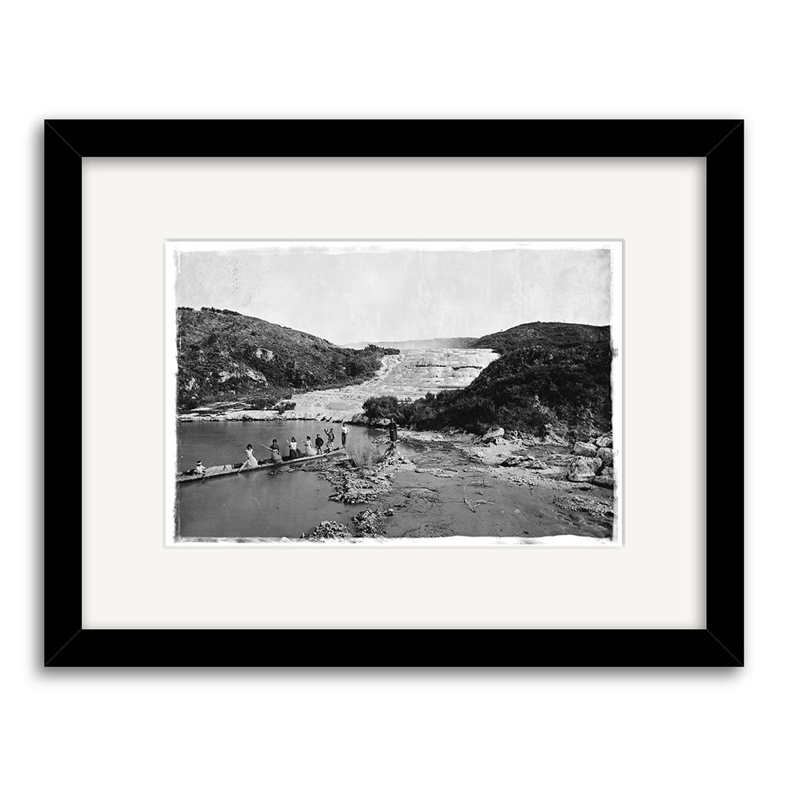 Archival-quality black and white photographic print, Pink Terraces 1879-1886 - HI064, shows several people on a log over a river amid layered rocks and hills. Presented in a black frame with white mat; captures New Zealand’s historical landscape.