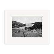 This archival-quality historical print, Pink Terraces 1879-1886 - HI064 from New Zealand, shows a group crossing a wooden bridge over water with rocky hills and a large geothermal terrace in the background. Wide white mat included.