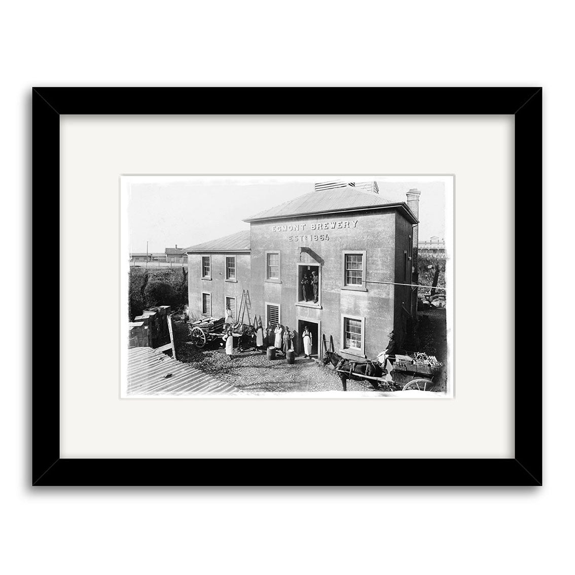 Black-and-white photo titled Overlook New Plymouth 1875 - HI068 of a two-story brick building with people, barrels, and carts outside. This historical photographic print is made in New Zealand and framed with a black border.