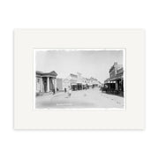 Black and white historical photographic print Alfred Street 1911 - HI070 shows a wide street in a historic New Zealand town with wooden buildings, shops, signs, and people. Mounted with a white archival-quality mat.