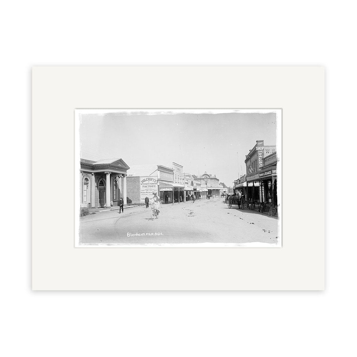 Black and white historical photographic print Alfred Street 1911 - HI070 shows a wide street in a historic New Zealand town with wooden buildings, shops, signs, and people. Mounted with a white archival-quality mat.