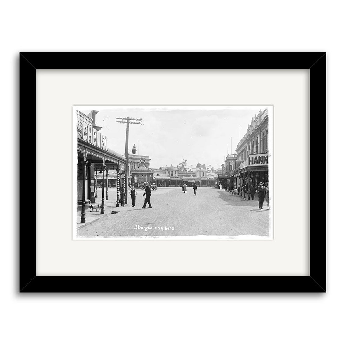 Archival quality print of Market Place 1911 - HI073: black-and-white New Zealand street scene with people, shops, and visible CHEMIST and HANN signs. Framed with a white mat and black frame.