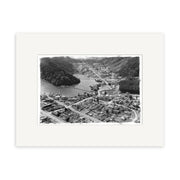 The Picton 1958 - HI082 Historical Photographic Print, a black and white aerial photo on archival matte paper, features a small town amid forested hills, water to the left, dense buildings in front, and comes displayed with a white mat.
