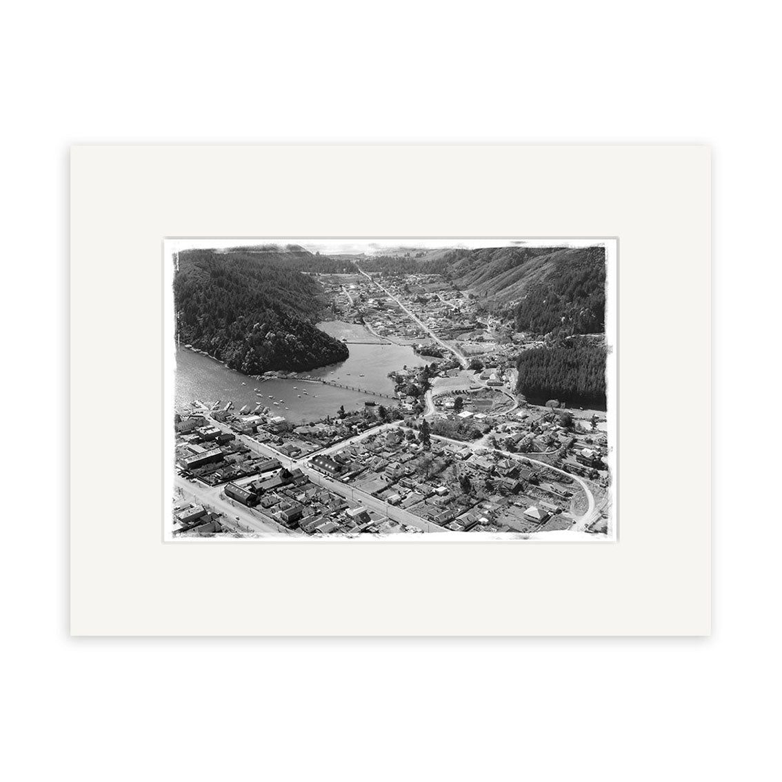 The Picton 1958 - HI082 Historical Photographic Print, a black and white aerial photo on archival matte paper, features a small town amid forested hills, water to the left, dense buildings in front, and comes displayed with a white mat.