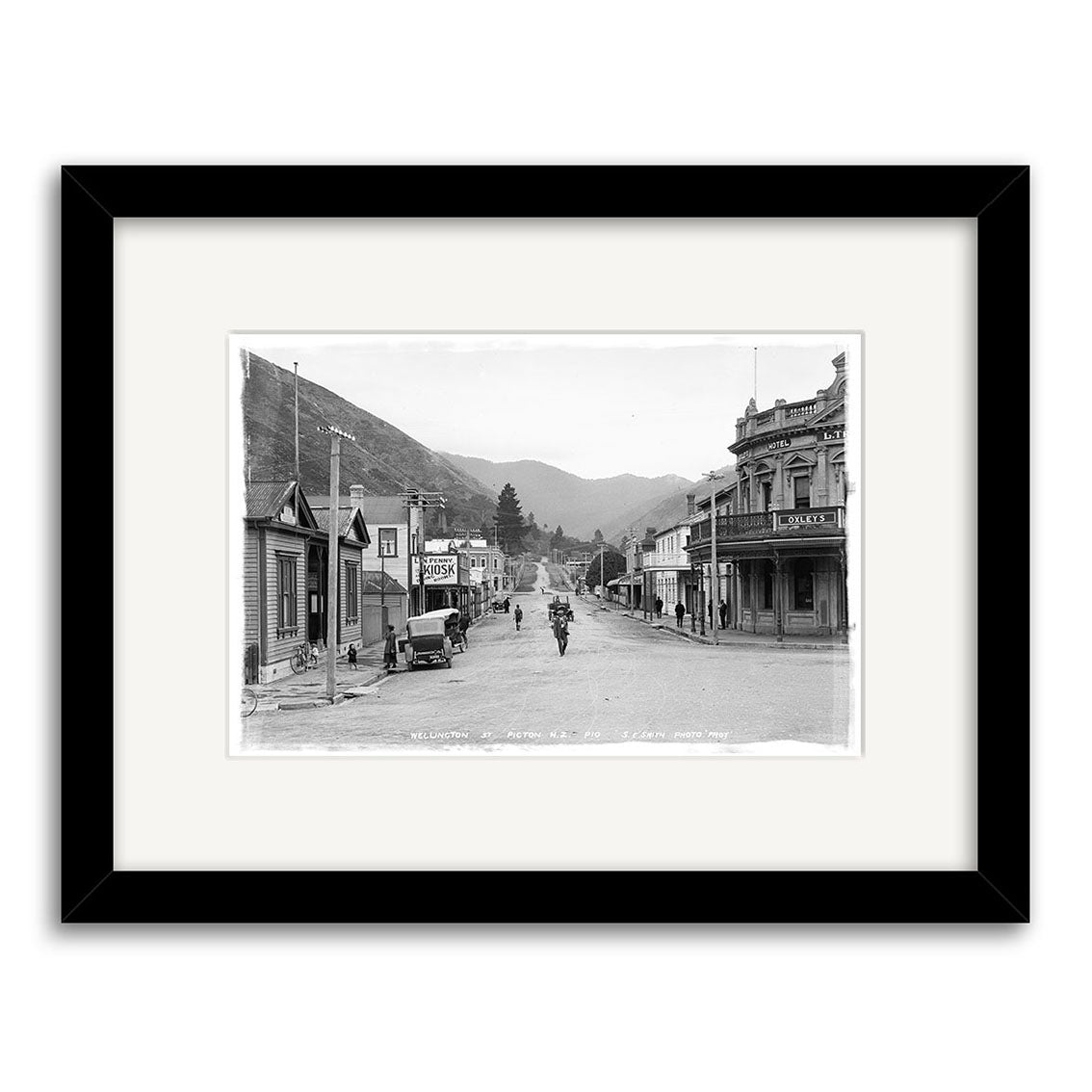 The Wellington St Picton 1910 - HI083 is a black and white historical photographic print of a small-town street with people, a horse-drawn carriage, and mountains, printed on archival matte paper and framed with a white mat and black frame.