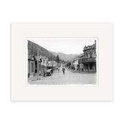 Black-and-white photo titled Wellington St Picton 1910 - HI083 shows a street in a small New Zealand town lined with wooden buildings, storefronts, several people, a horse-drawn carriage, and hills in the background.