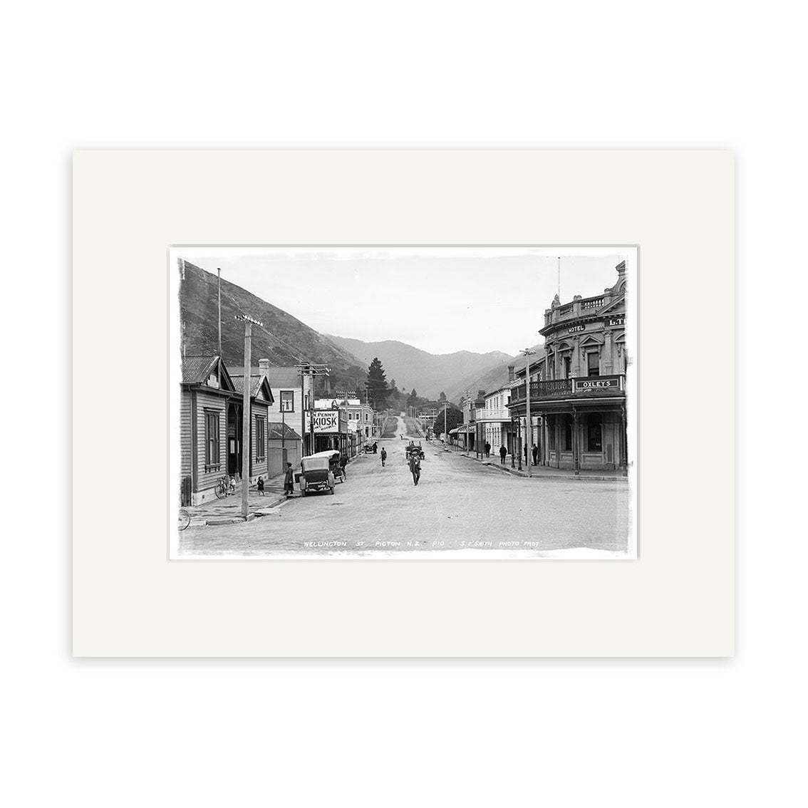 Black-and-white photo titled Wellington St Picton 1910 - HI083 shows a street in a small New Zealand town lined with wooden buildings, storefronts, several people, a horse-drawn carriage, and hills in the background.