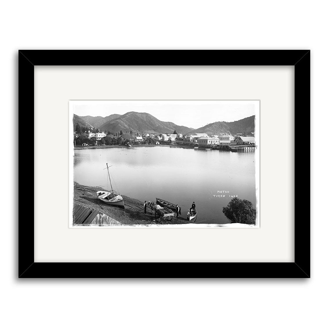 The Picton 1890 - HI084 is a black and white framed historical photographic print depicting a calm lake with boats, waterfront buildings, distant mountains, and people by the shore. Printed on archival quality matte paper.