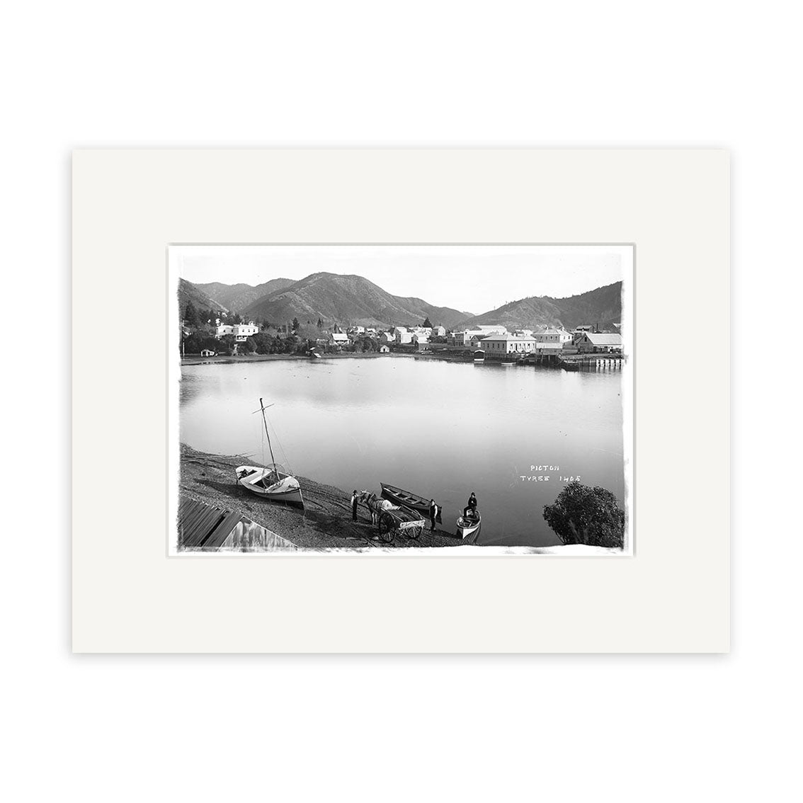 Picton 1890 - HI084: Black and white historical photographic print of a lakeside town with boats, people, and hillside buildings. Framed with a white mat on archival-quality matte paper for lasting beauty.