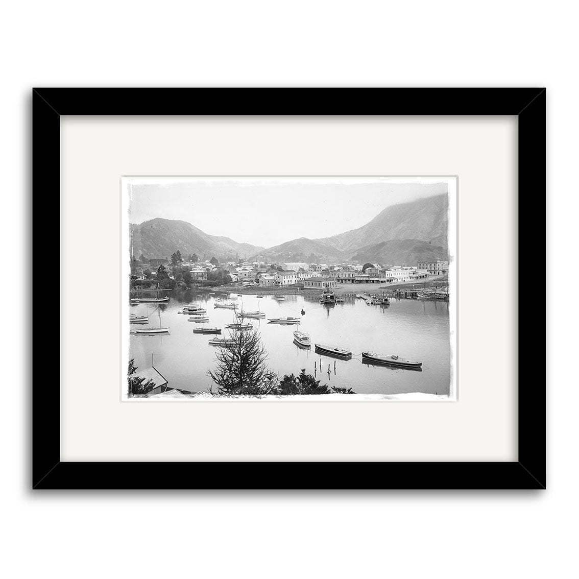 Picton Harbour 1920 - HI085: Black and white historical photo print of a coastal New Zealand town with boats in the harbor and mountains behind. Archival matte paper, white mat, black frame.
