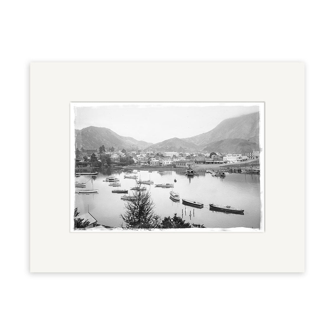Black and white photograph titled Picton Harbour 1920 - HI085 shows small boats, shoreline buildings, trees, and distant mountains. This historical print is matted with a white border on archival-quality matte paper.