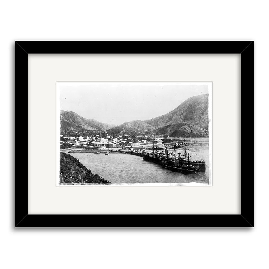 Black-and-white historical print, Picton Township 1897 - HI086: a New Zealand coastal town with shorefront buildings, mountains, and a docked ship. Presented in a black frame with an archival quality white mat.