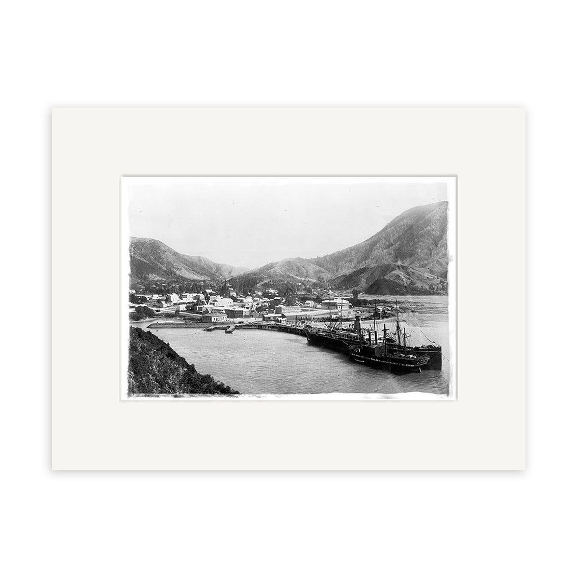 Archival-quality black and white photographic print, Picton Township 1897 - HI086, depicts a New Zealand coastal town with shoreline buildings, a docked ship, and hills; professionally framed with a white mat.