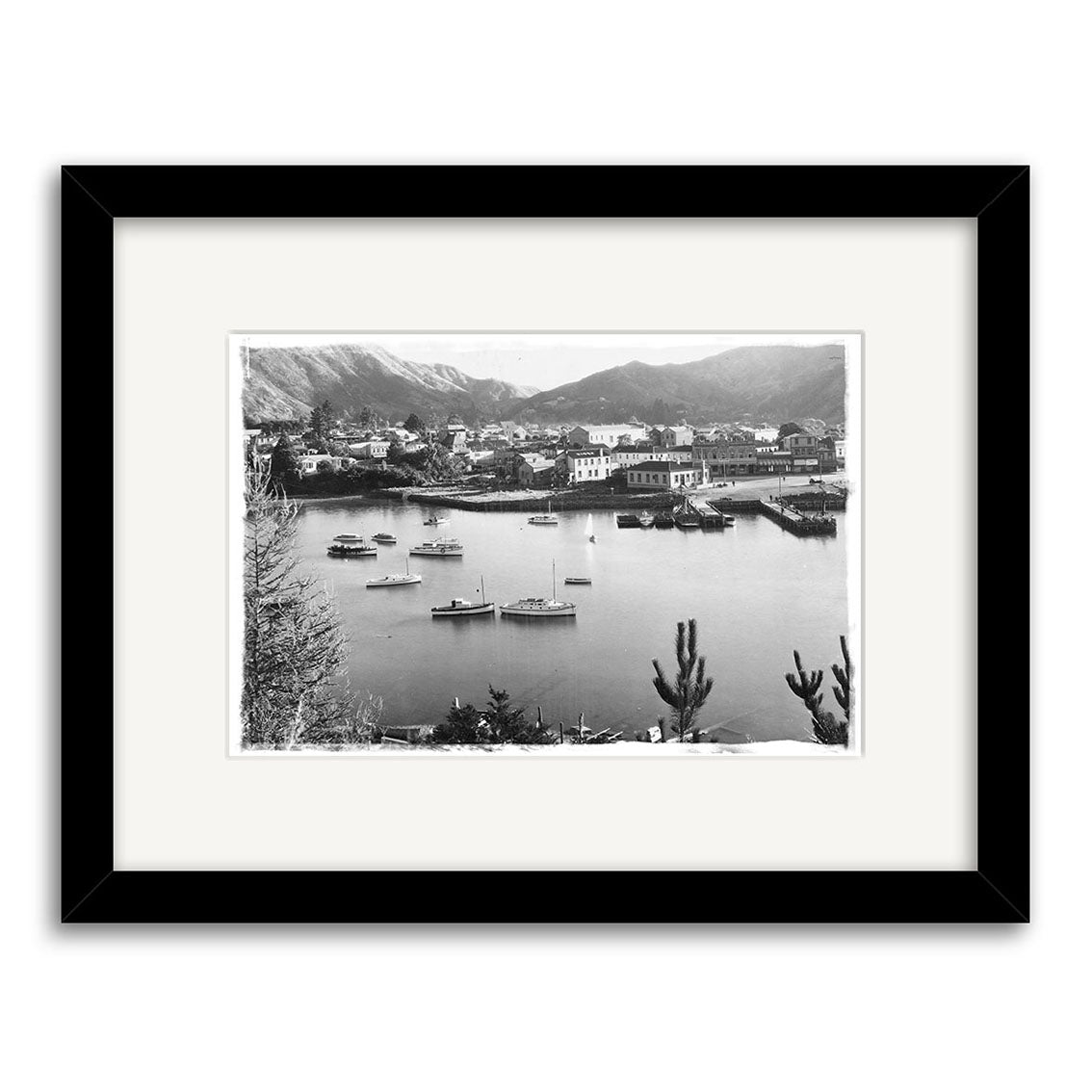 Archival quality black and white photo of Picton Harbour, 1920s, with boats near the shore, buildings, and mountains. Displayed in a black frame with white mat. A striking historical photographic print from New Zealand - Picton Harbour 1920s - HI087.
