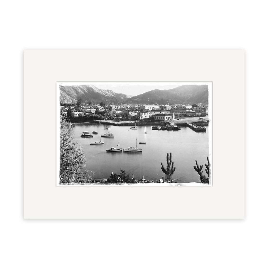 Picton Harbour 1920s - HI087: Black and white photo of a New Zealand harbor with boats, a small town, and mountains. Trees frame the foreground. Archival-quality print matted with a white border.