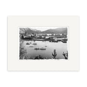 Picton Harbour 1920s - HI087: Black and white photo of a New Zealand harbor with boats, a small town, and mountains. Trees frame the foreground. Archival-quality print matted with a white border.