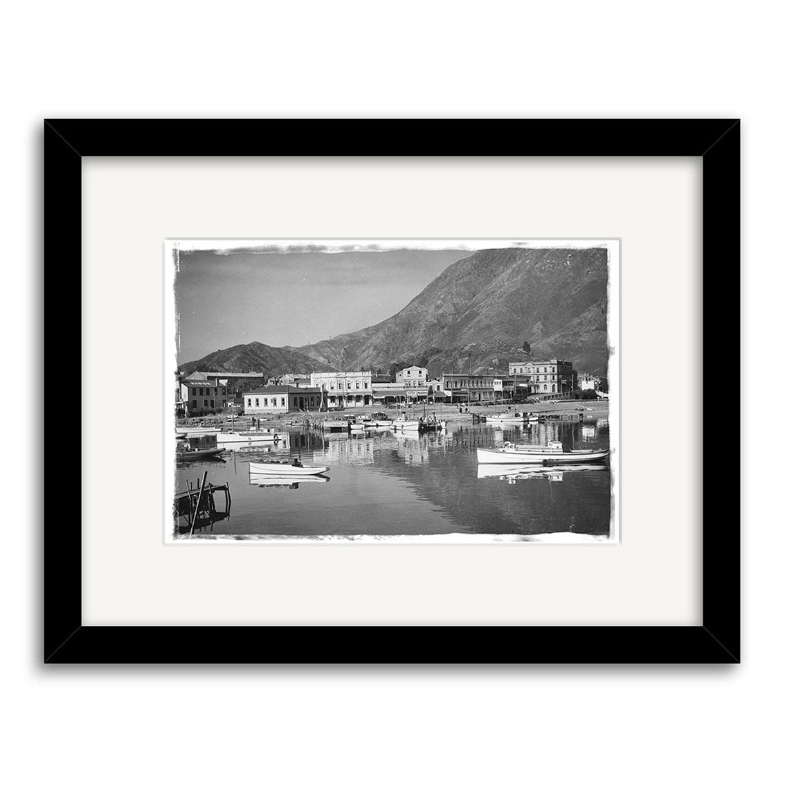 Picton Harbour 1930 - HI088: Black and white photo print of a harbor with small boats, waterfront buildings, and a distant mountain. Framed with a white mat and black frame for classic, timeless style.