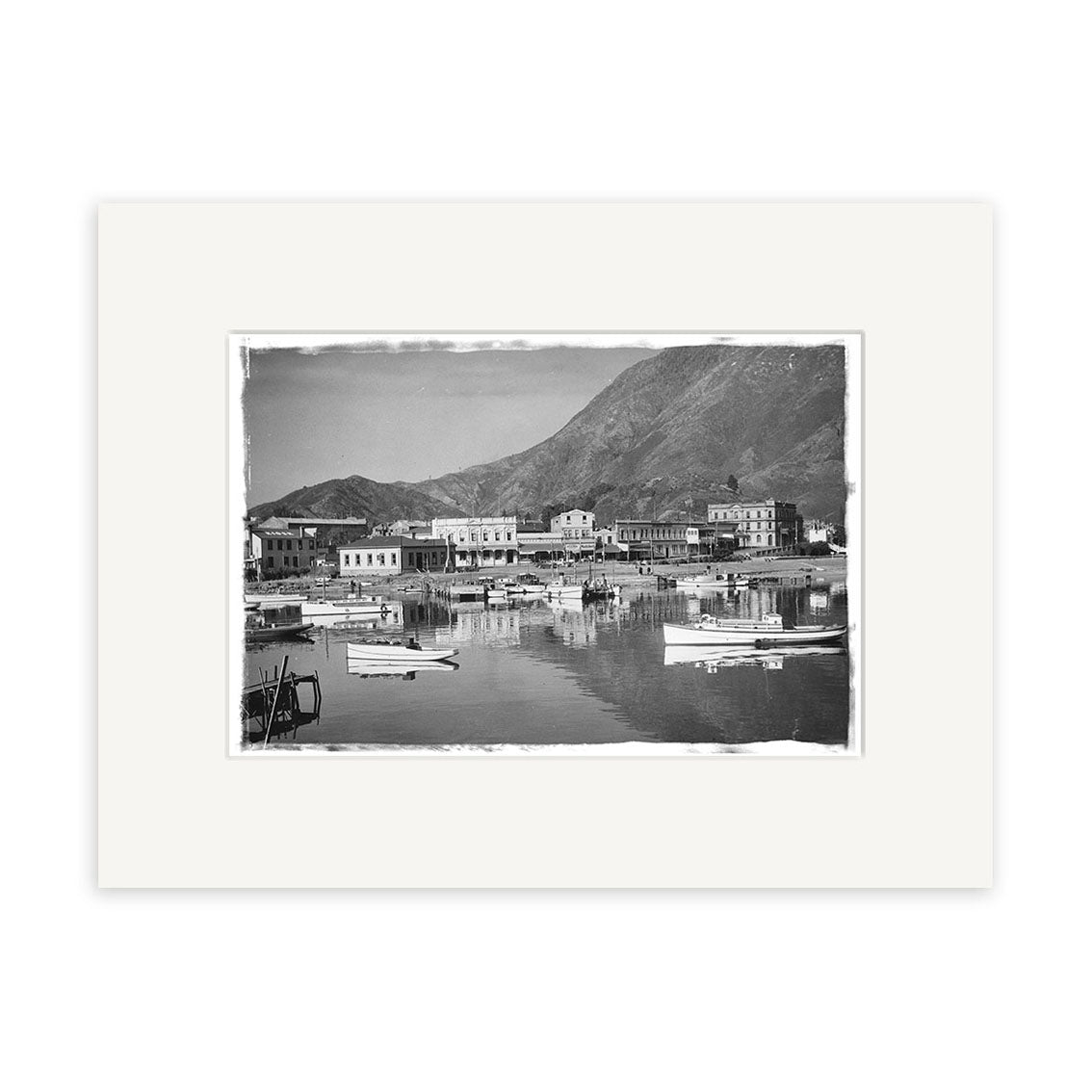 Picton Harbour 1930 - HI088: Black and white photo of a waterfront town with boats, buildings on shore, and mountain backdrop, centered with a white border on archival matte paper.