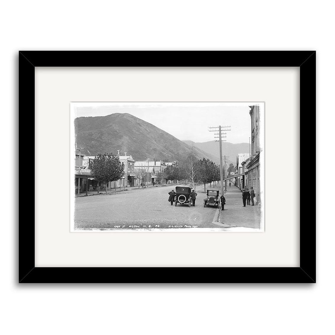 Archival-quality black & white photo print: “High Street Picton 1910 - HI091” shows a quiet NZ street with vintage cars, people, and hills. Framed with a white mat and black frame.