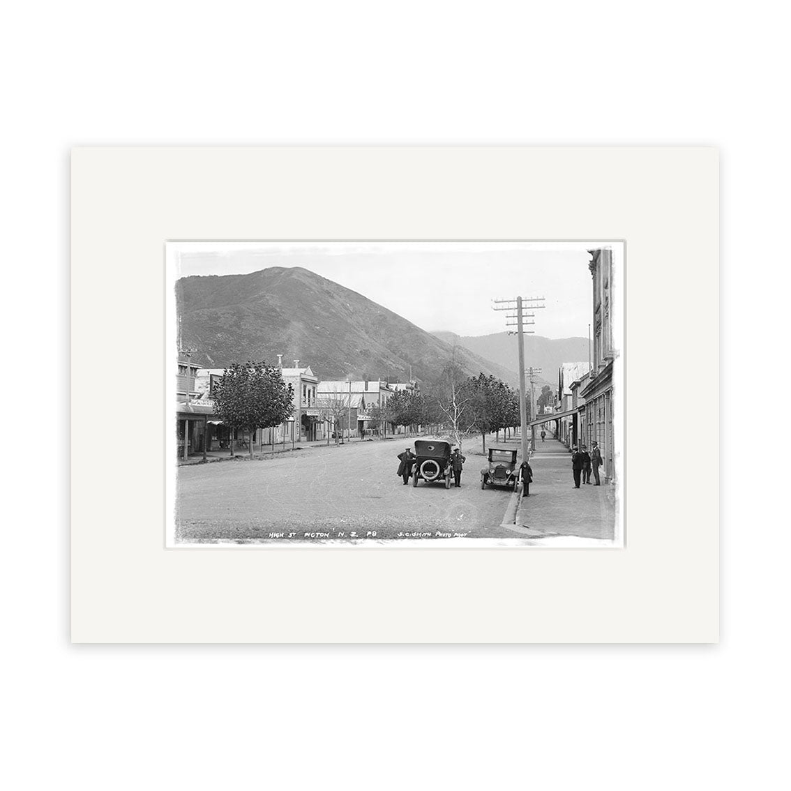 Archival quality Historical Photographic Print: High Street Picton 1910 - HI091 shows a black and white view of a wide street in small-town New Zealand, early 1900s cars parked along buildings, people nearby, and a mountain backdrop.