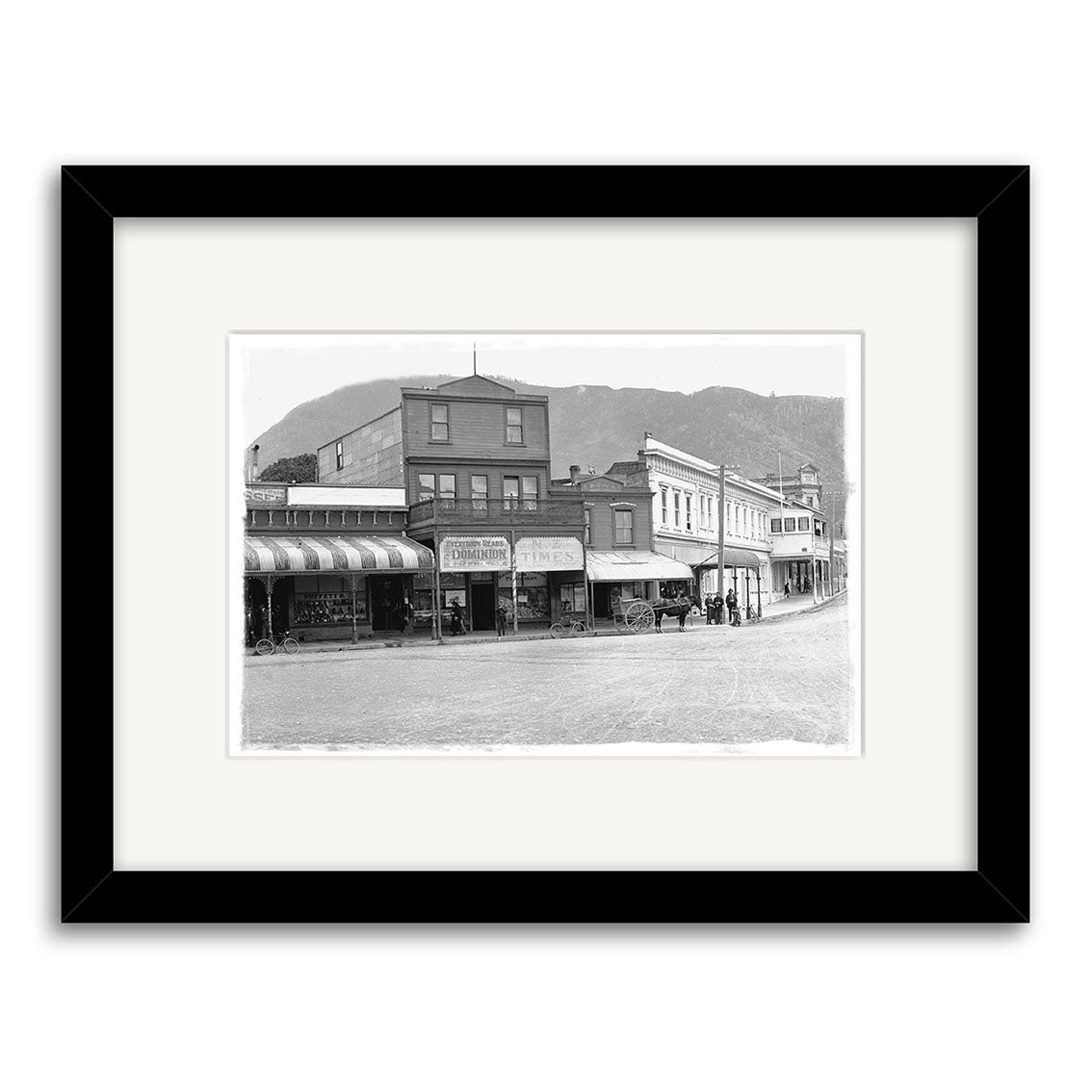 Black-and-white photo London Quay Picton 1910 - HI093: A historic small-town street with storefronts, horse-drawn carriages, people on the sidewalk, and mountains behind. Historical Photographic Print made in New Zealand, framed in black.