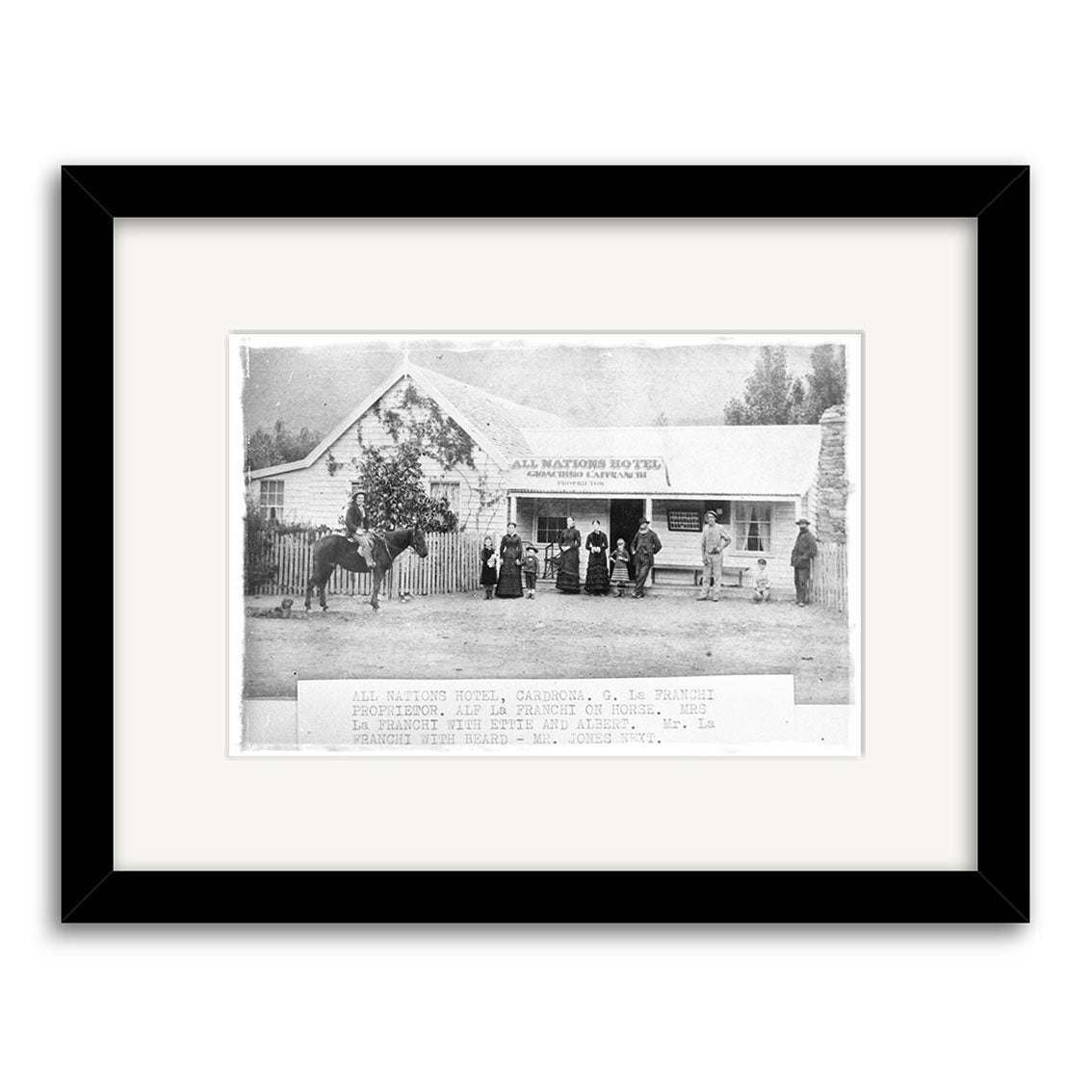 A black-and-white historical photographic print, “Cardrona 1870 - HI094,” shows a group before the All Nations Hotel in New Zealand. Framed with a white mat and black border, it’s perfect for archival-quality display.