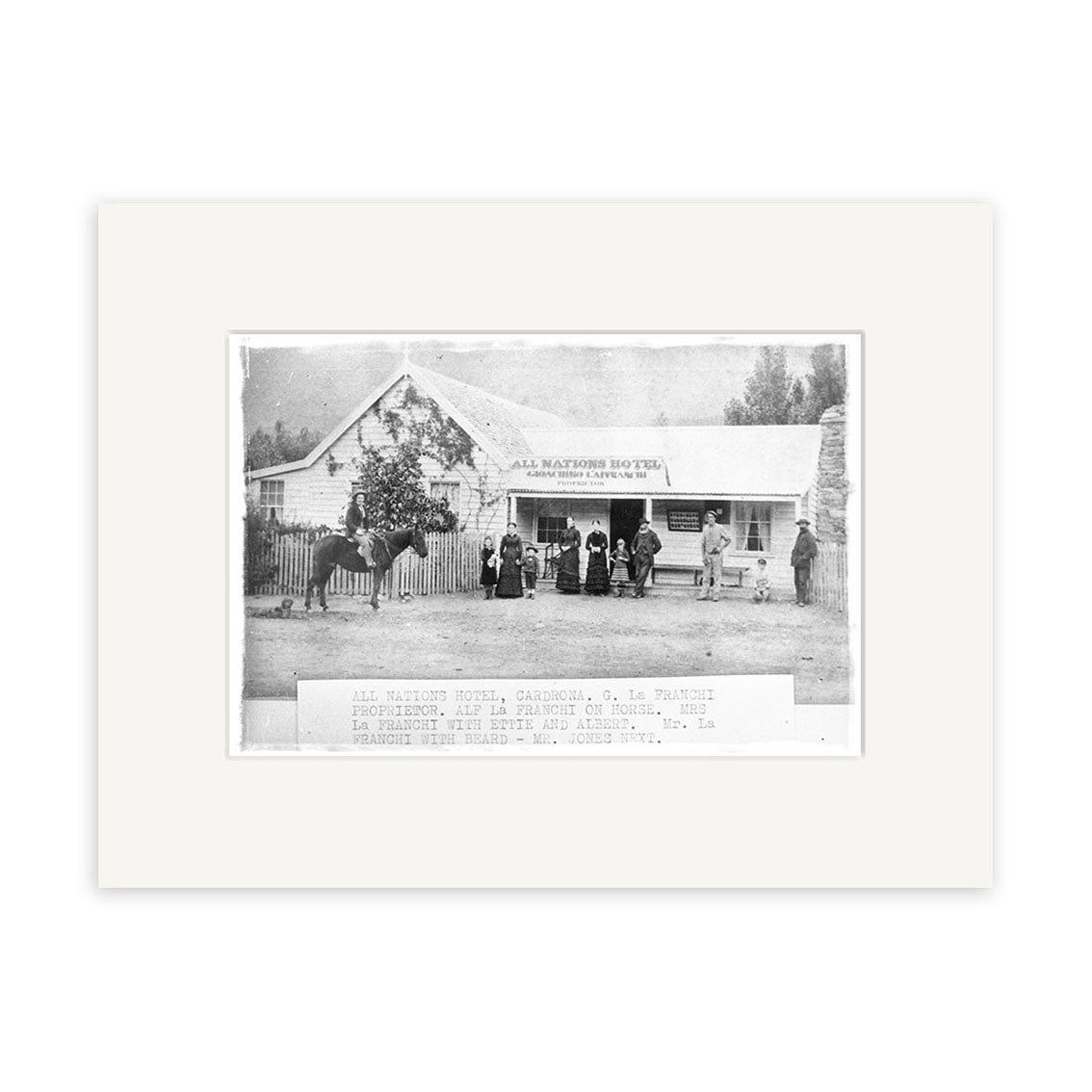 Black and white archival quality photographic print titled Cardrona 1870 - HI094, showing a group outside the All Nations Hotel in New Zealand, featuring a pitched roof, picket fence, and tree to the left; label included.