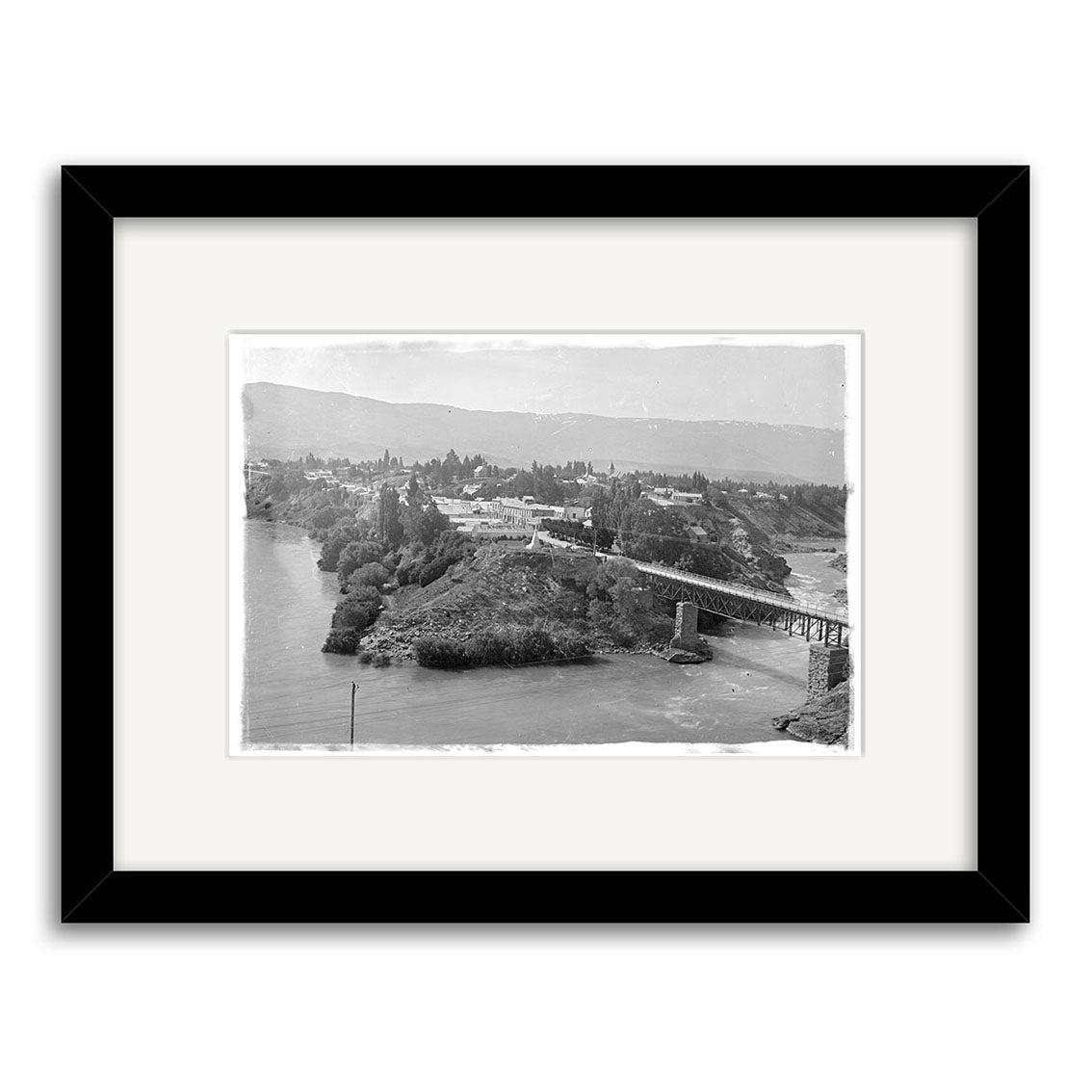Cromwell Clutha River 1926 - HI095: A black and white historical photographic print of a riverside town with bridge, trees, and hills. Framed in black, printed on archival matte paper in New Zealand.