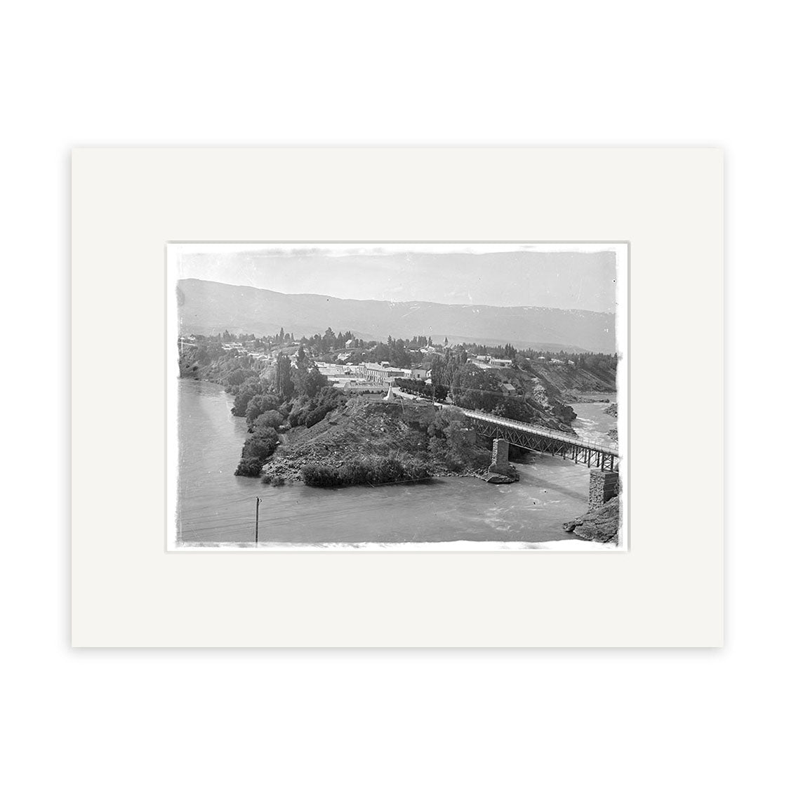 The Cromwell Clutha River 1926 - HI095 is a black and white historical photographic print of a small riverside town with a bridge, hills, and trees. Printed on archival matte paper and beautifully framed with a wide white mat.
