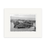 The Cromwell Clutha River 1926 - HI095 is a black and white historical photographic print of a small riverside town with a bridge, hills, and trees. Printed on archival matte paper and beautifully framed with a wide white mat.