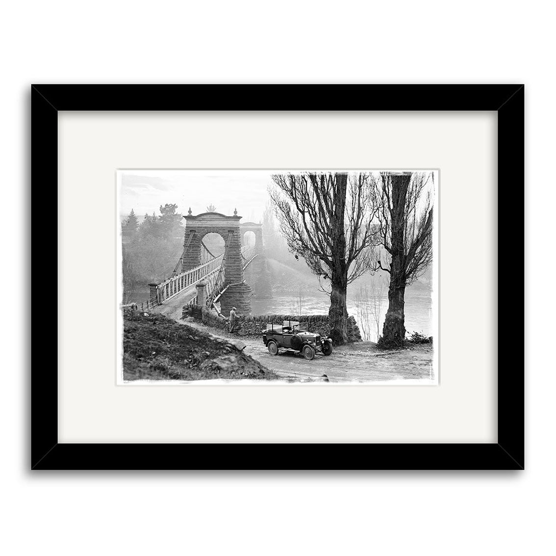 The Otago Central Bridge 1930 - HI098 is a black and white photo of a vintage car by tall trees and a suspension bridge, framed in black with a white mat. Made in New Zealand on archival matte paper for lasting beauty.