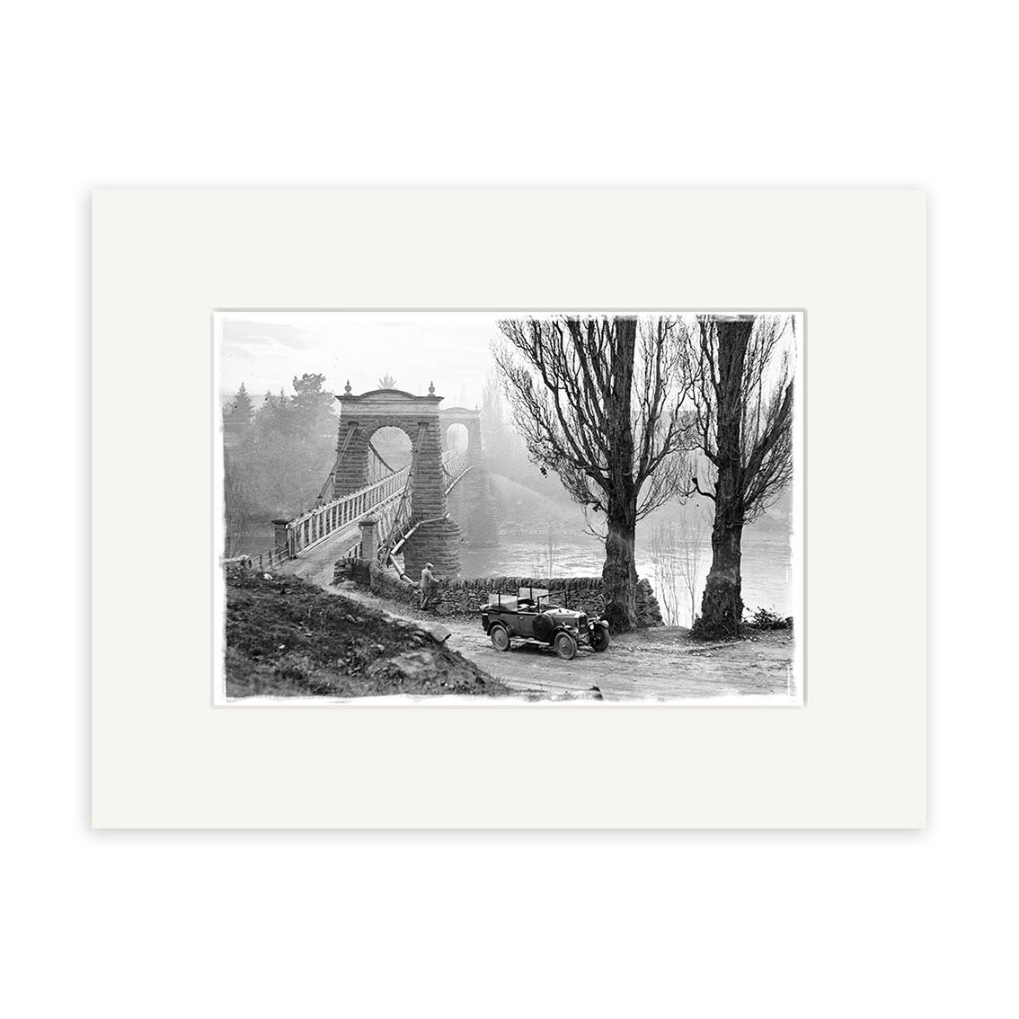 A vintage black-and-white photo titled Otago Central Bridge 1930 - HI098 shows an old car by a road near tall trees and the stone suspension bridge over water in New Zealand, with dense foliage and mist in the background.