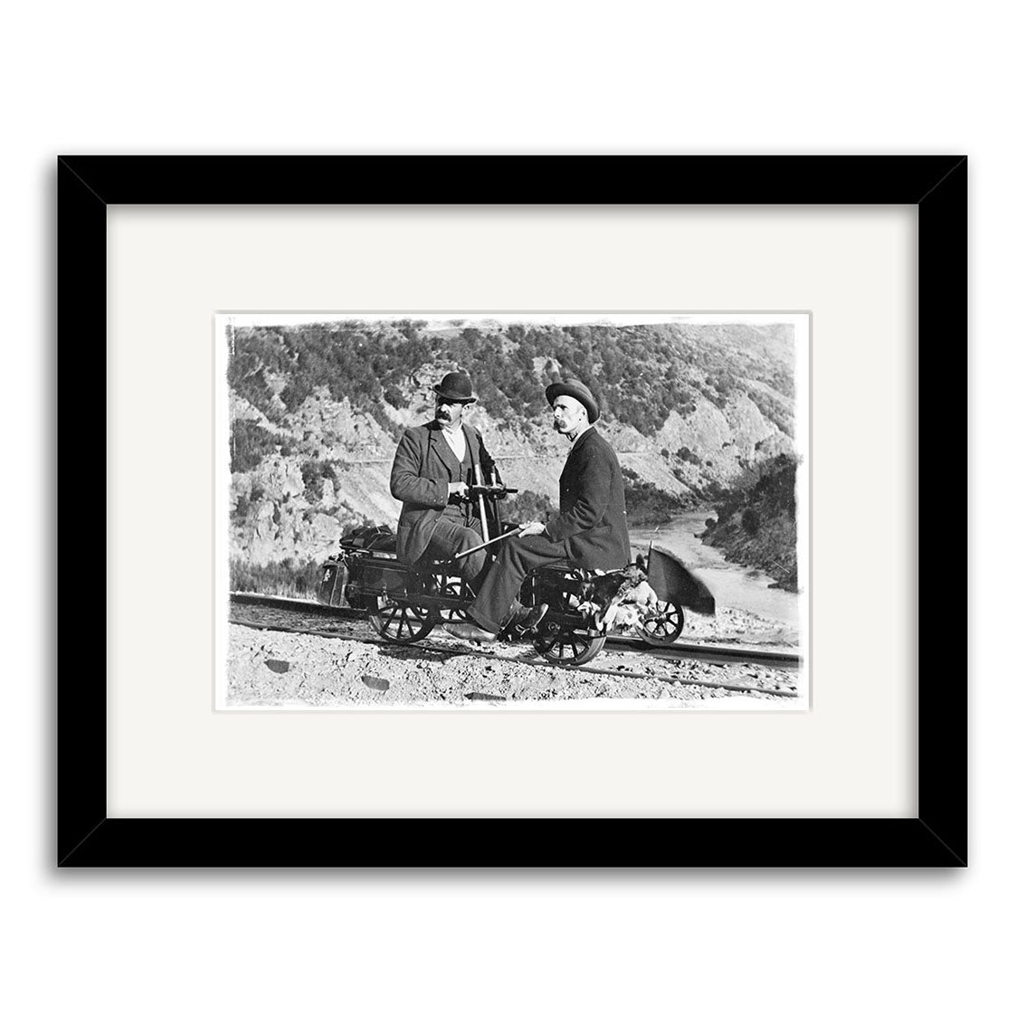 Otago 1900 - HI099: A black-and-white historical photo from New Zealand shows two people in early 20th-century attire on a small rail vehicle in a hilly landscape. Framed with a white mat and black frame.