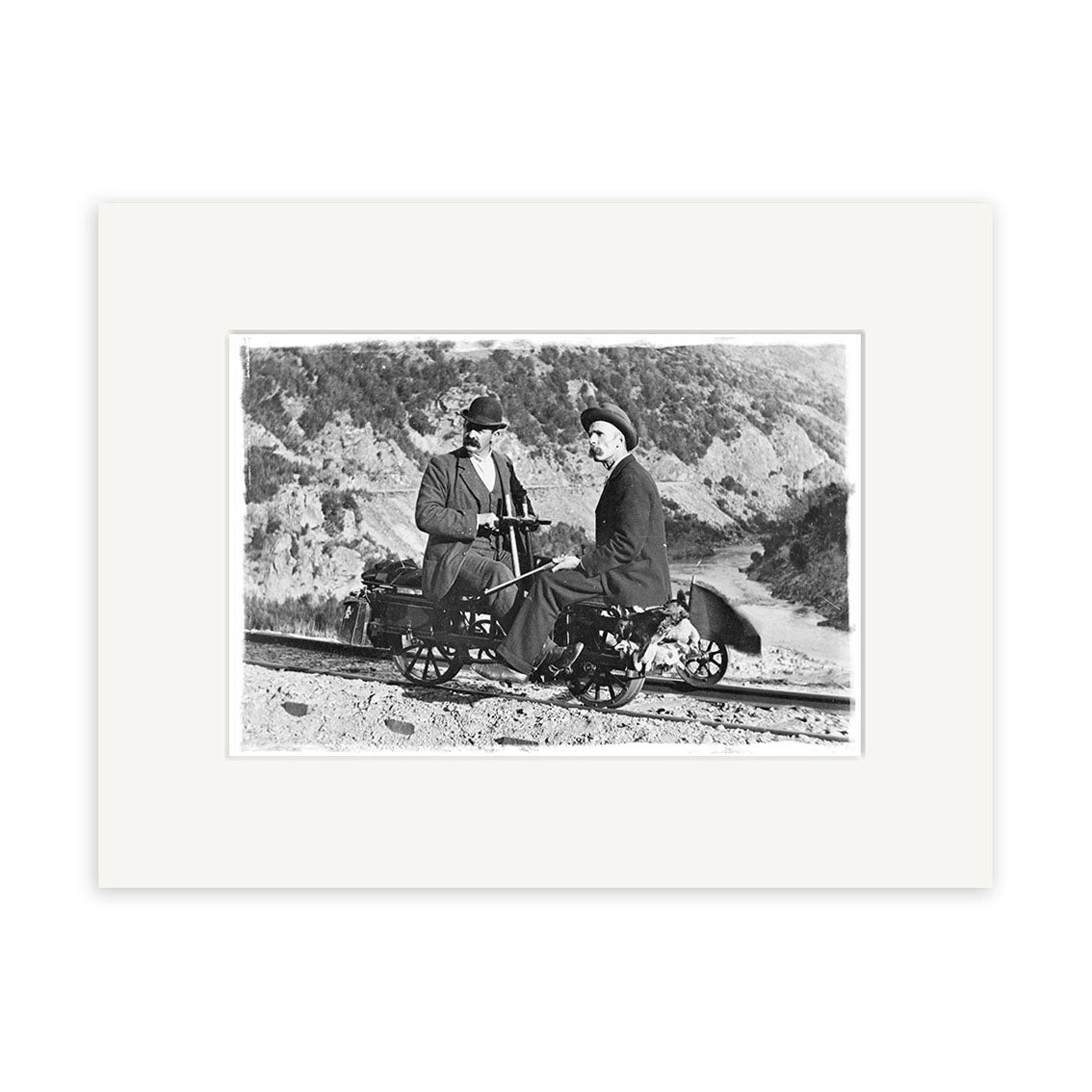 Otago 1900 - HI099 is a historical photographic print showing two people in early 20th-century dress on a handcar along railroad tracks, with hills behind them, made in New Zealand on archival matte paper.